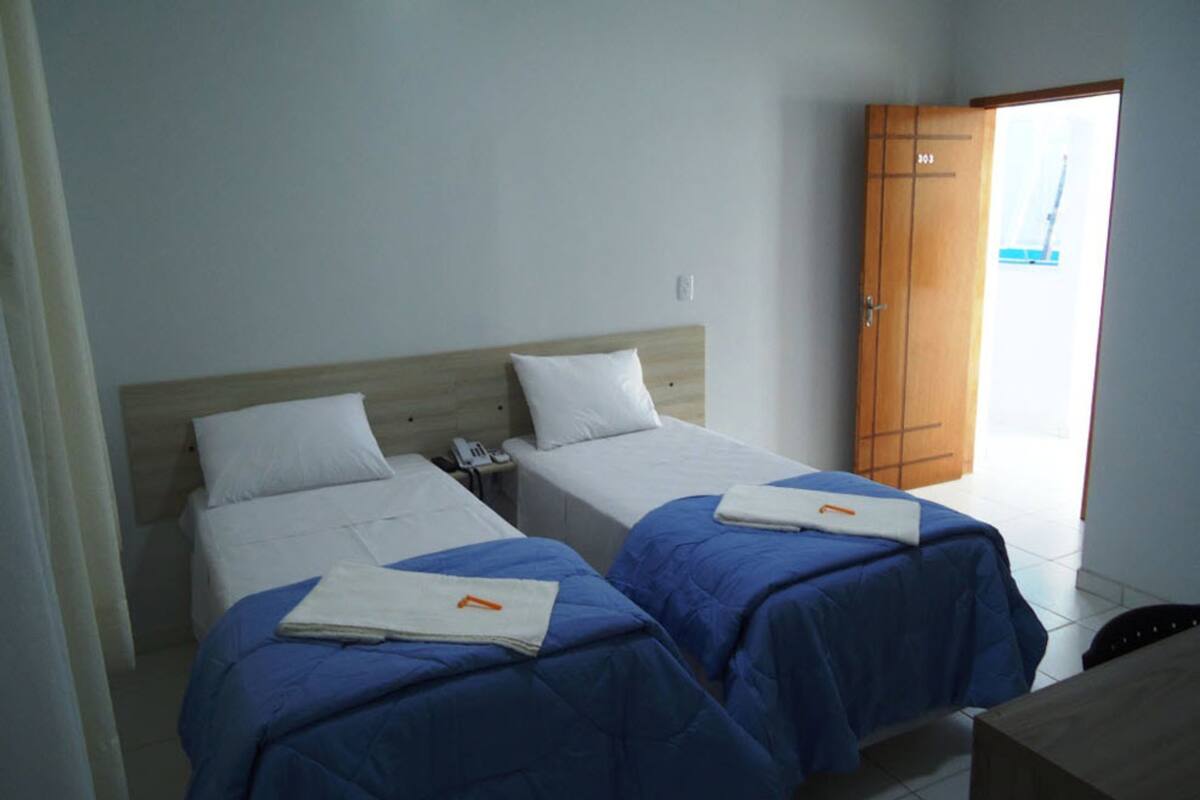 Ett hotellrum med två separata sängar, var och en med ett blått överkast och vita kuddar. I rummet finns en garderob i trä och ett litet bord.