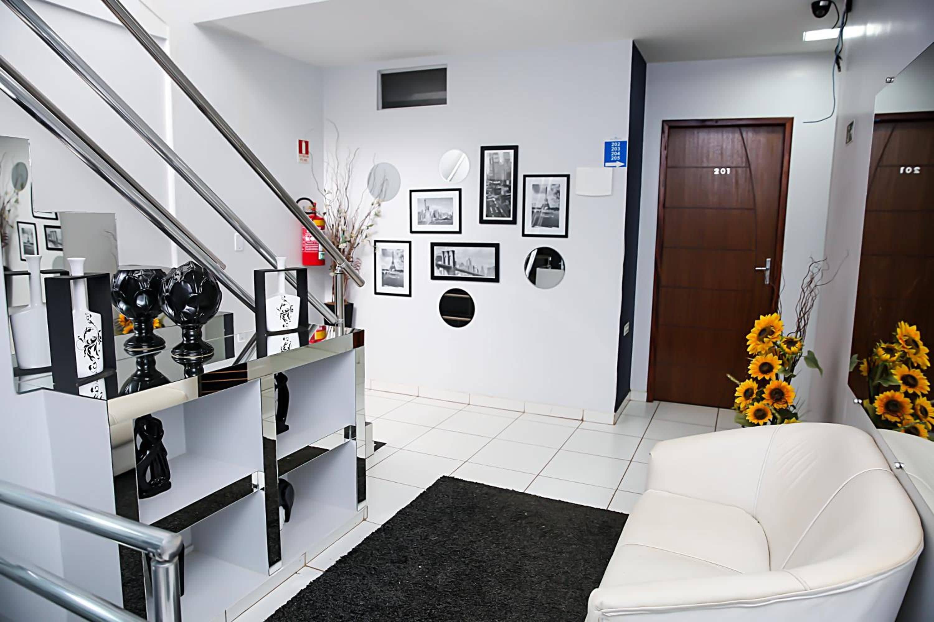 Um hall de hotel com escadas, balcão de recepção, quadros nas paredes, uma porta com números de quartos e um sofá com girassóis.