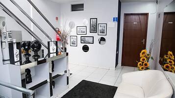 Um hall de hotel com escadas, balcão de recepção, quadros nas paredes, uma porta com números de quartos e um sofá com girassóis.