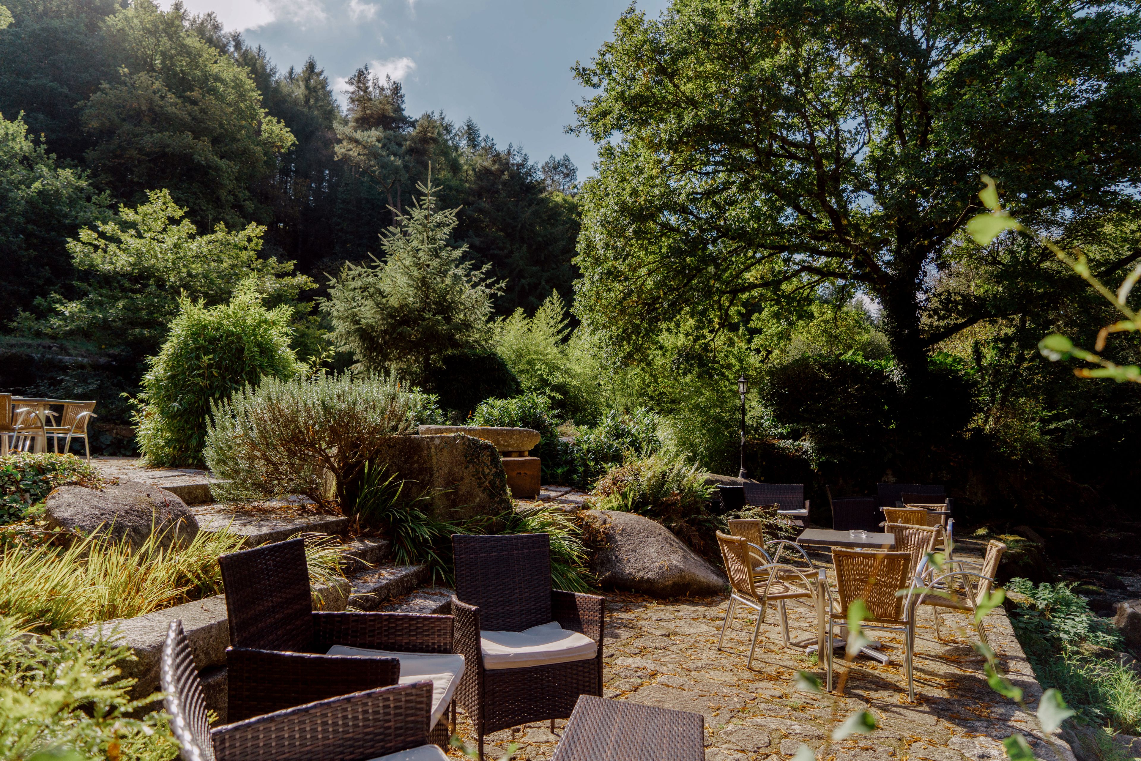A patio area with wicker furniture and a stone floor, surrounded by lush greenery and trees.