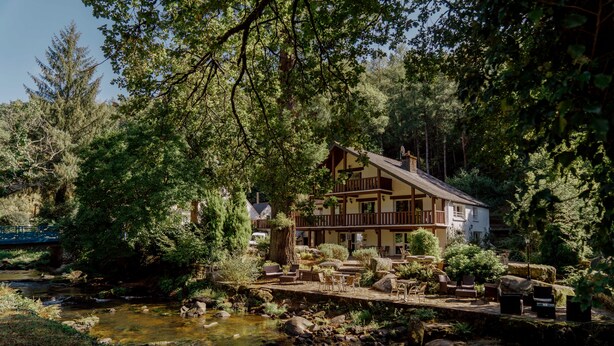 A lodge with a deck overlooking a stream, surrounded by dense forest.
