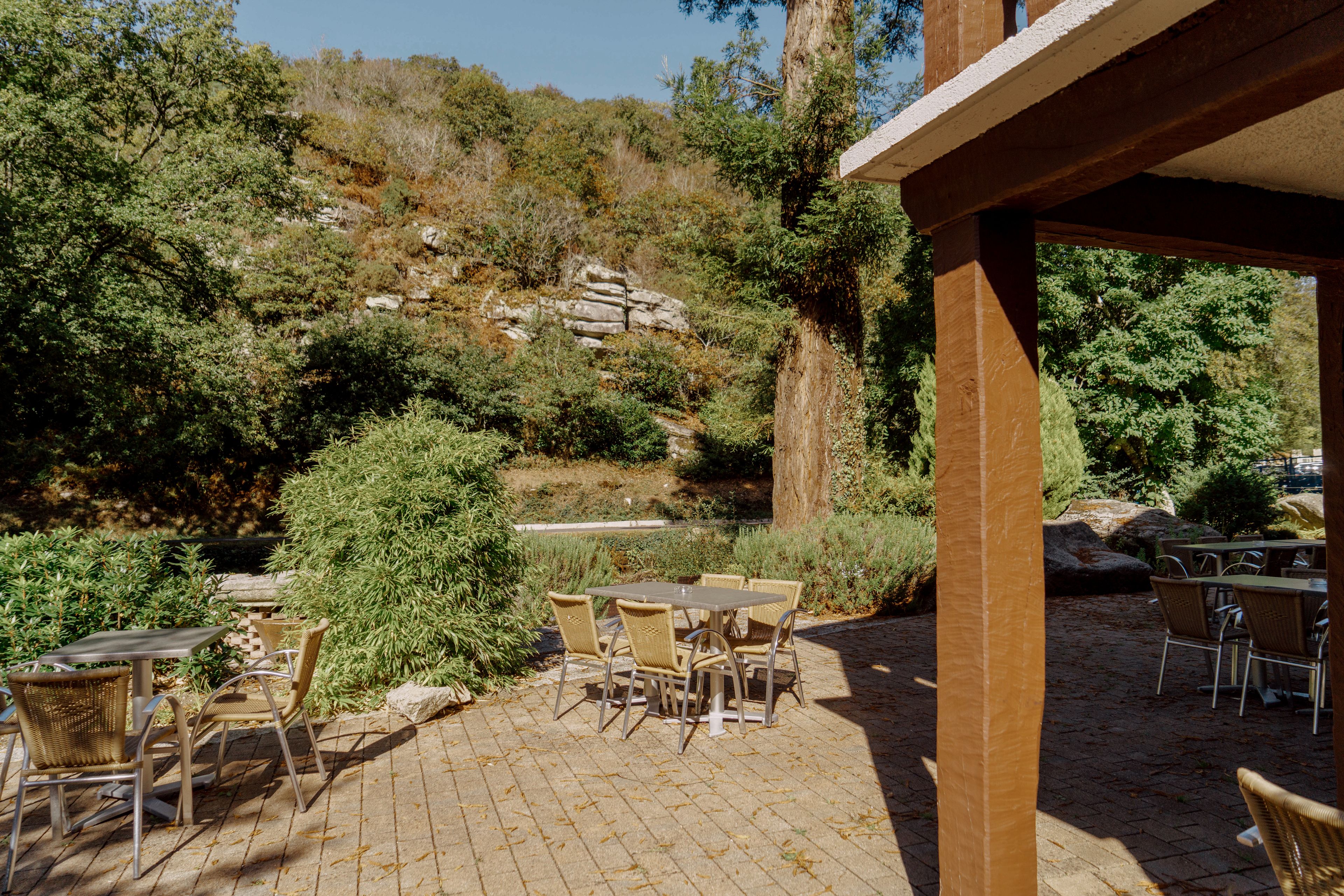 Un patio al aire libre con mesas y sillas, rodeado de vegetación natural y una ladera rocosa.