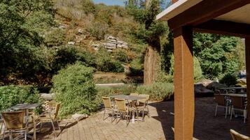 Un patio al aire libre con mesas y sillas, rodeado de vegetación natural y una ladera rocosa.
