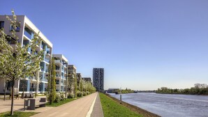 A riverside walkway with modern buildings, a tall skyscraper, and a clear sky. - Plenus Riverloft (Bremen)