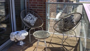 A balcony with two wicker chairs, one with a geometric pillow, and a metal table with a silver bowl of white flowers. - Plenus Riverloft (Bremen)