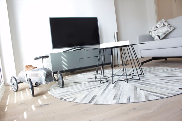 A modern living room with a flat-screen TV, a grey sofa, a patterned rug, and a vintage car model. - Plenus Riverloft (Bremen)