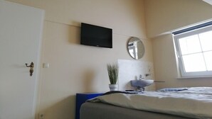 Un dormitorio con cama, televisor montado en la pared, espejo redondo, planta en maceta y ventana.