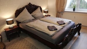 A neatly made bed with a wooden headboard, two bedside tables with lamps, a window with curtains, and a potted plant on the windowsill. - Nijole Apartment (Winterberg)