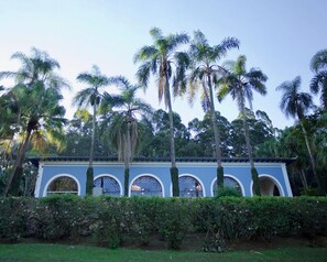 A building with a blue facade and arched windows, surrounded by lush greenery and tall palm trees. - Residencial Sênior Club Leger (São Paulo)