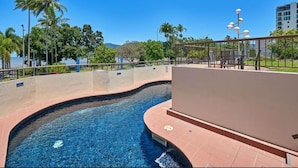 A swimming pool with a curved design, surrounded by a low wall and a railing. There are palm trees and a clear blue sky in the background. - Cairns Ocean View Apartment in Aquarius (Cairns)