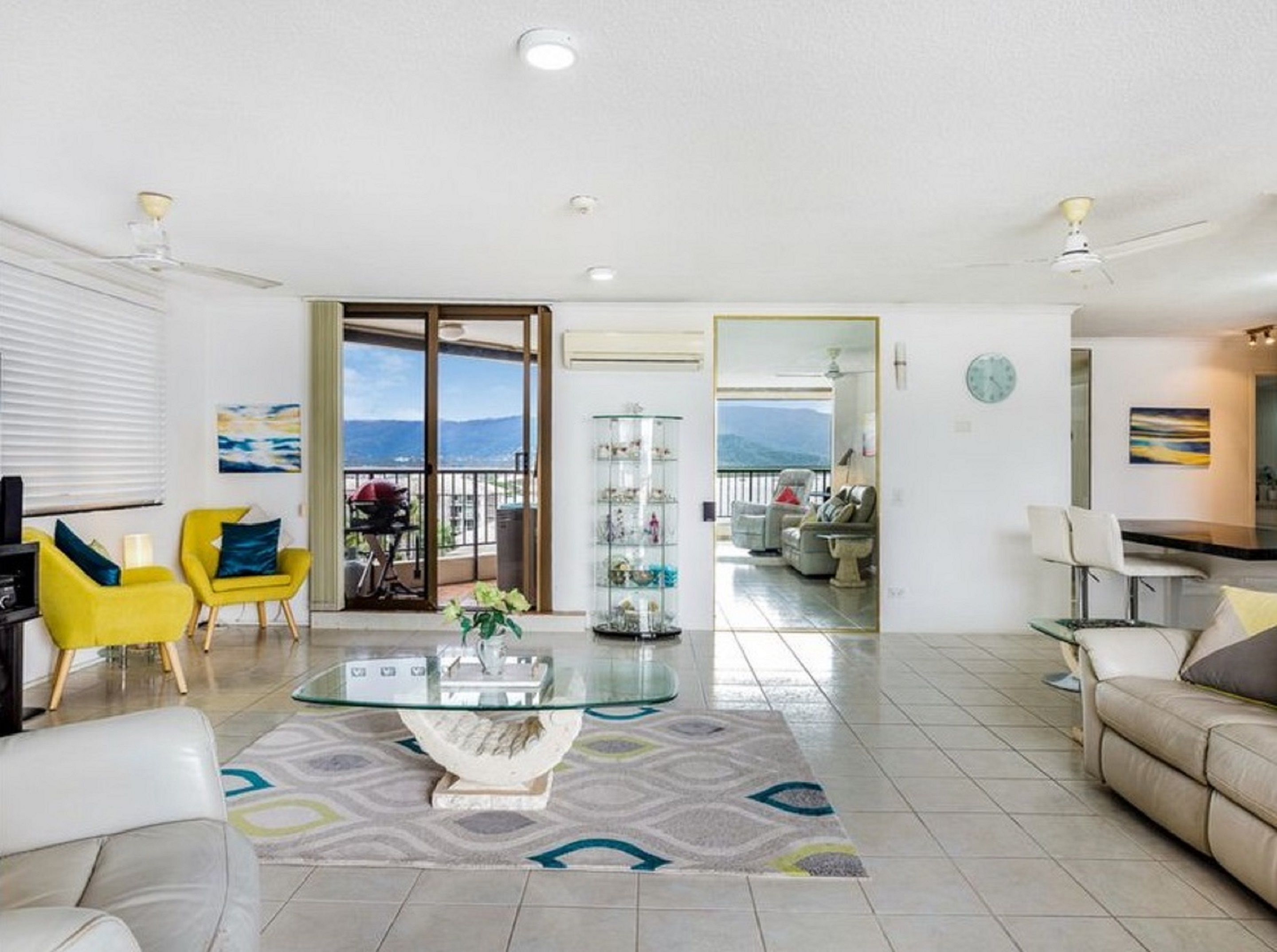 A spacious living room with a glass coffee table, yellow chairs, a beige sofa, and a view of a balcony with a mountain in the background.