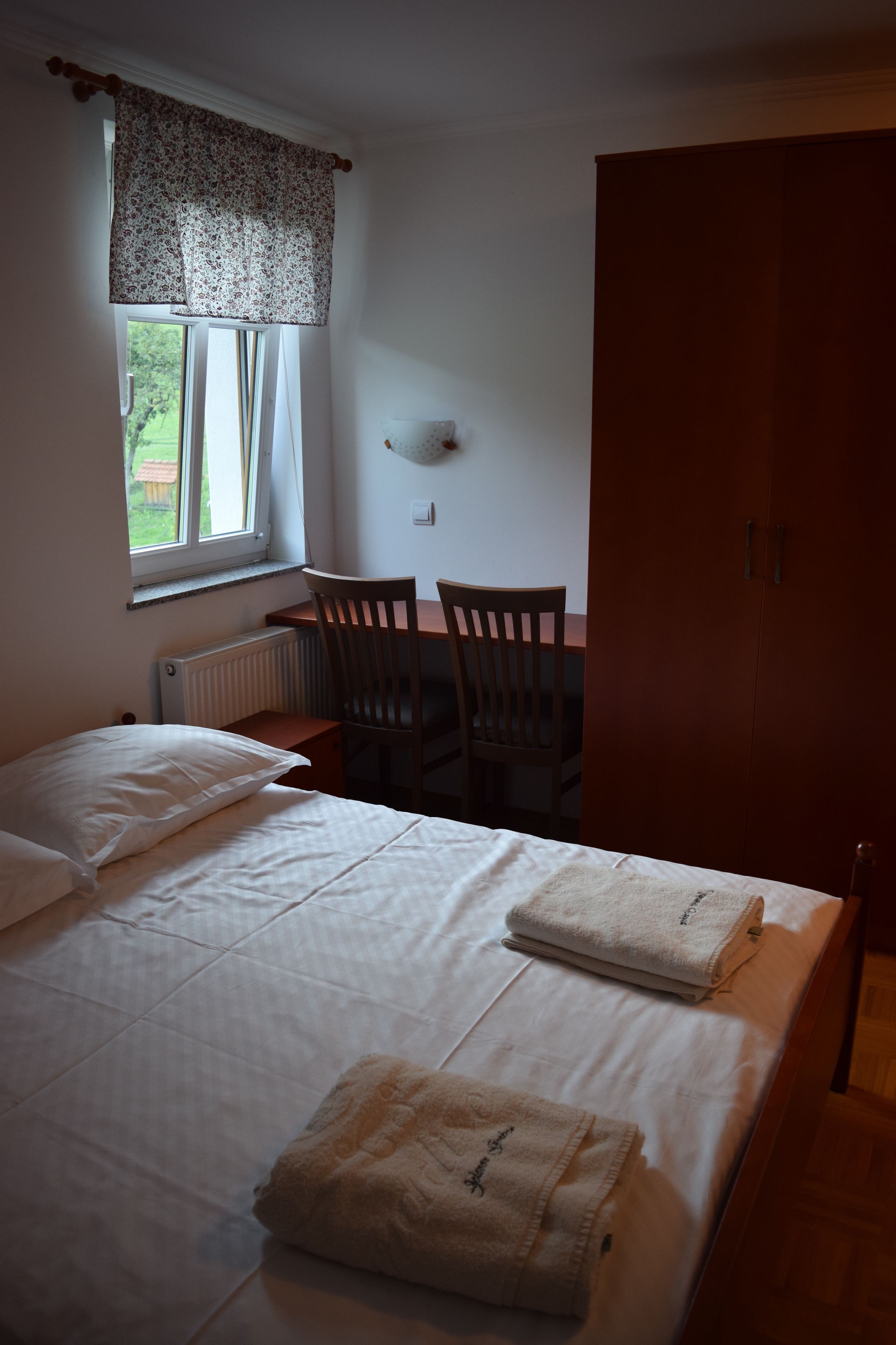 Een slaapkamer met een bed, twee stoelen, een tafel, een radiator en een raam met gordijnen.