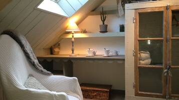 Una habitación acogedora con techo inclinado, un sillón blanco, una mesa con lámpara y un aparador con puertas de vidrio.