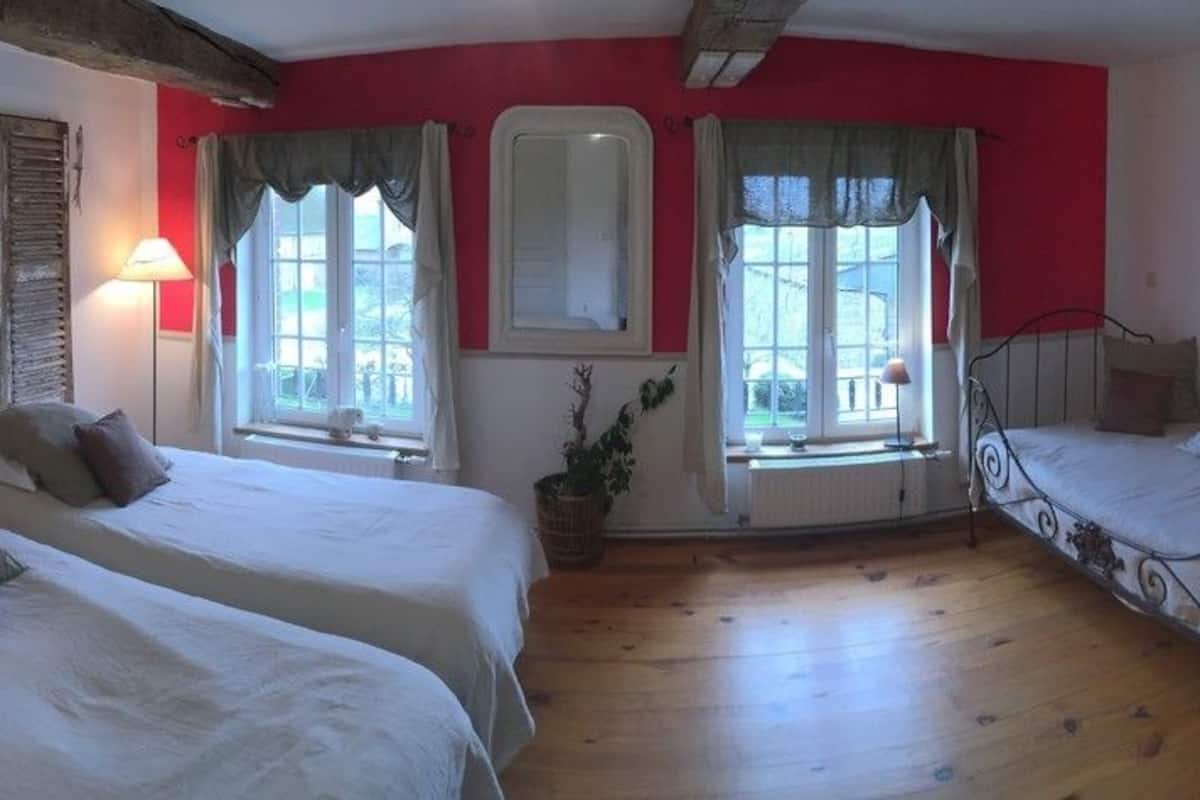 Ein Zimmer mit zwei Betten, Holzboden, einer roten Akzentwand und einem Fenster mit Vorhängen.