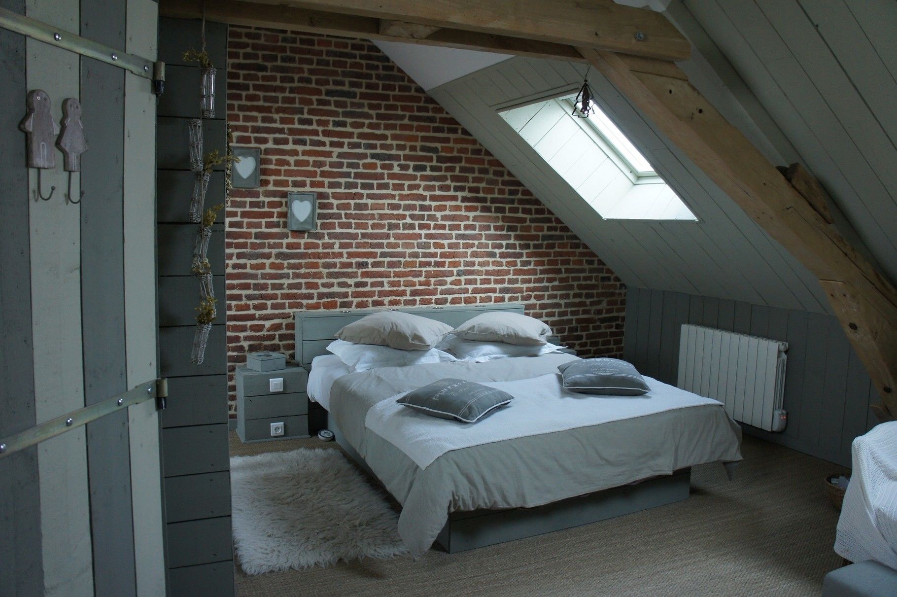 Une chambre située dans un espace de type « loft », avec un plafond en pente, des murs en briques apparentes, un puits de lumière, un lit avec des oreillers, une table de chevet et un radiateur.