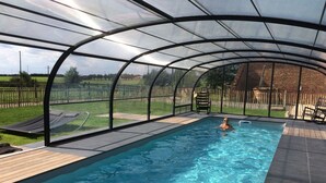 Una piscina techada con techo transparente, rodeada por una terraza de madera y un jardín cercado.