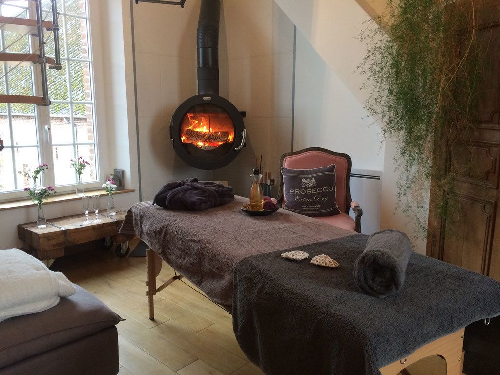 Une pièce chaleureuse avec un poêle à bois, une table de massage et une fenêtre ornée de fleurs.