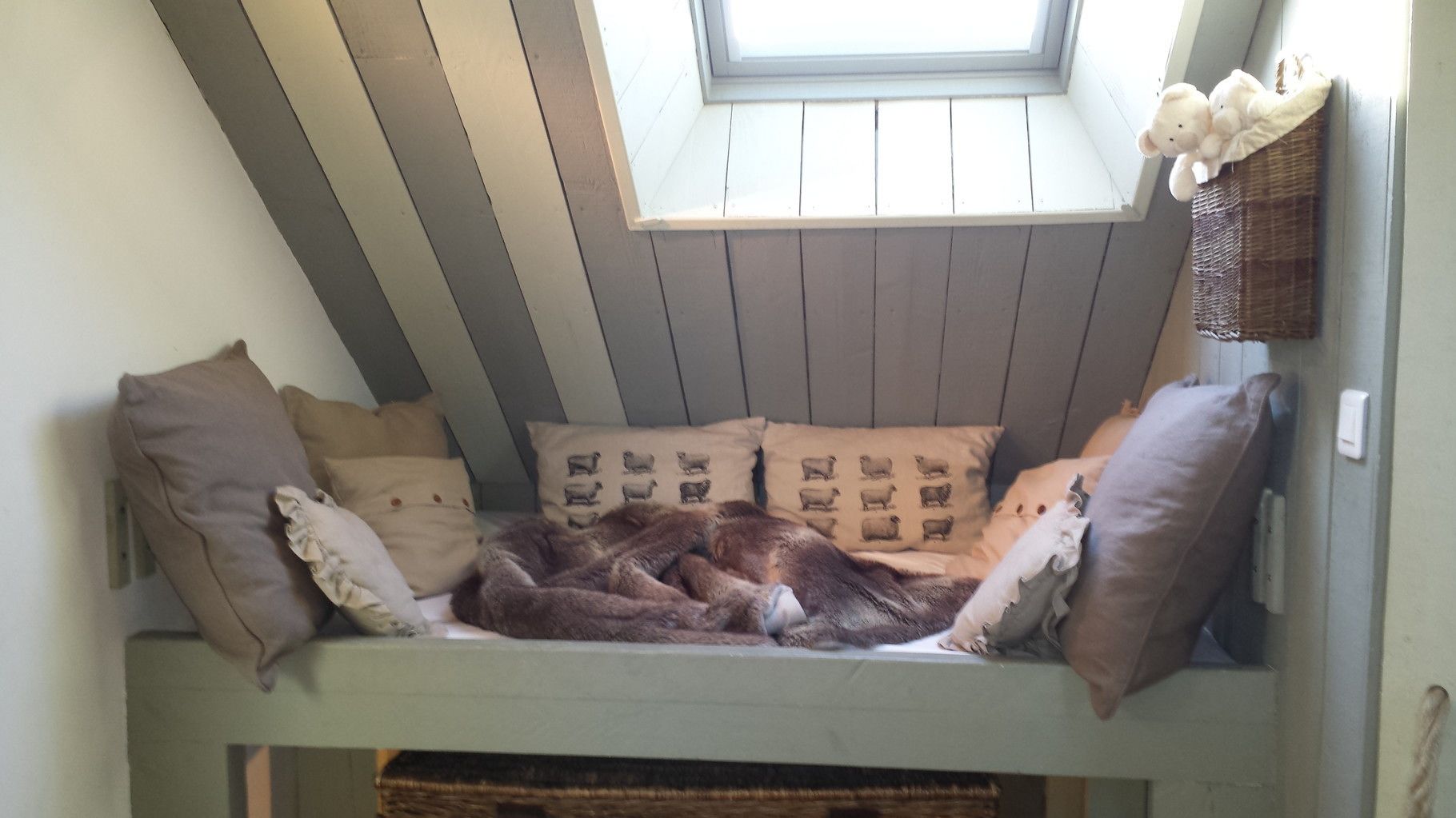A cozy loft-style bed with a sloped ceiling and a skylight, featuring a variety of pillows and a woven basket.