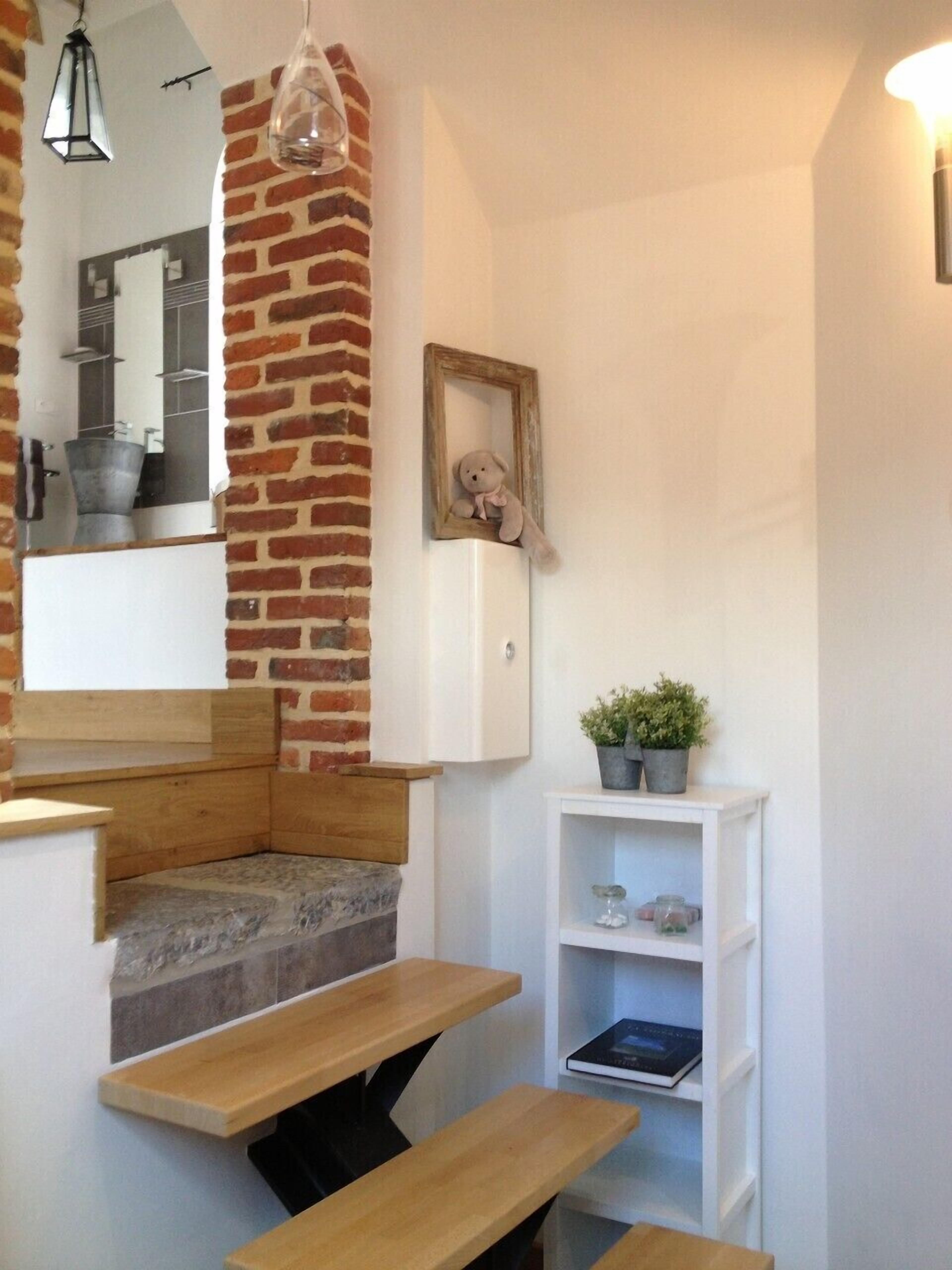 A cozy corner with a brick accent wall, a wooden bench, a small shelf with plants, and a mirror reflecting a bathroom area.