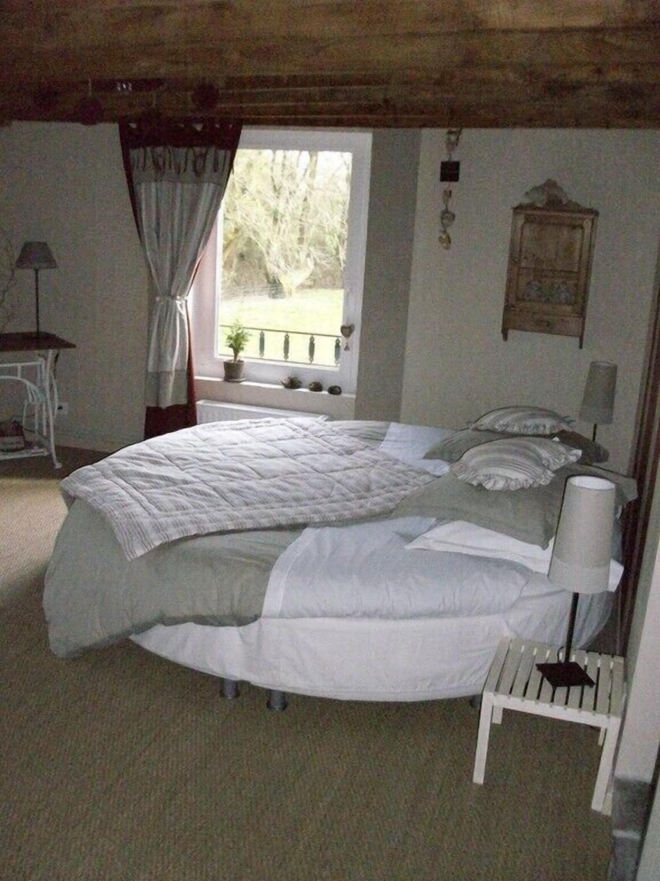 A bedroom with a large bed, a small table, a chair, a window with curtains, and a wall-mounted shelf.