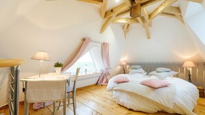 Un dormitorio con techo de madera, una cama con ropa de cama blanca y almohadas rosadas, un escritorio con lámpara y una ventana con cortinas.