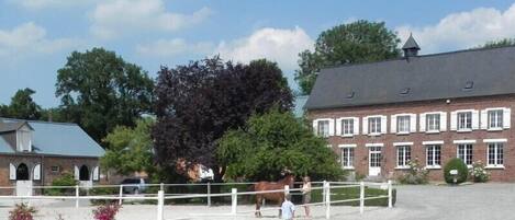Un establo con un caballo y una persona, rodeado por una cerca blanca, y un edificio de ladrillo al fondo.