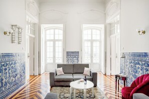 A room with a grey sofa, a white coffee table, and a red chair. There are two large windows with white frames and a blue patterned wall behind the sofa. - Trésor de Fern Apartment (Lisbon)