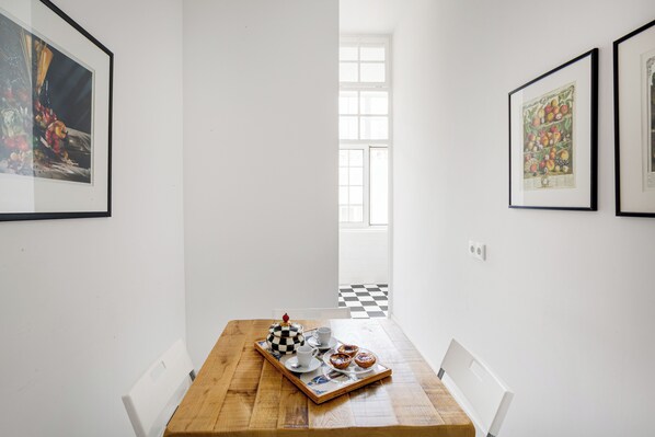 A dining area with a wooden table, white chairs, a tray with a teapot, cups, and pastries. There are framed pictures on the walls and a window with a checkered floor visible in the background. - Trésor de Fern Apartment (Lisbon)