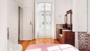 A bedroom with a wooden floor, a bed with a pink bedspread, a dresser with a mirror, and a door leading to a balcony. - Trésor de Fern Apartment (Lisbon)