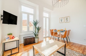 A modern living room with a wooden table, a TV mounted on the wall, and a dining area with orange chairs.