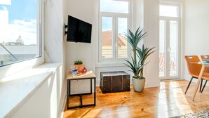 Une pièce lumineuse avec un plancher en bois, un téléviseur fixé au mur, une valise, une plante en pot et un coin repas avec des chaises.