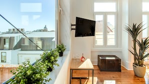Un salon moderne doté d’une grande fenêtre, d’un téléviseur à écran plat, d’une table en bois, d’une plante en pot et d’une valise ancienne.