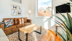 Un salon moderne avec un canapé en cuir marron, une table basse en bois et un tapis à motifs. On peut voir des œuvres d'art encadrées représentant des papillons accrochées au mur, une fenêtre donnant sur les toits et une plante en pot dans un coin.