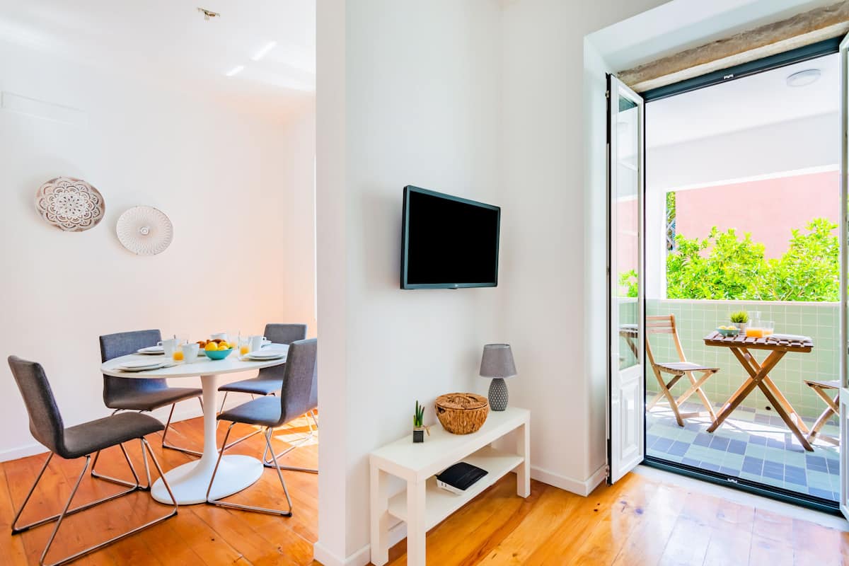 Uma área de jantar moderna com uma mesa redonda, quatro cadeiras, uma televisão de tela plana na parede e uma pequena mesinha de apoio com uma luminária e objetos decorativos.