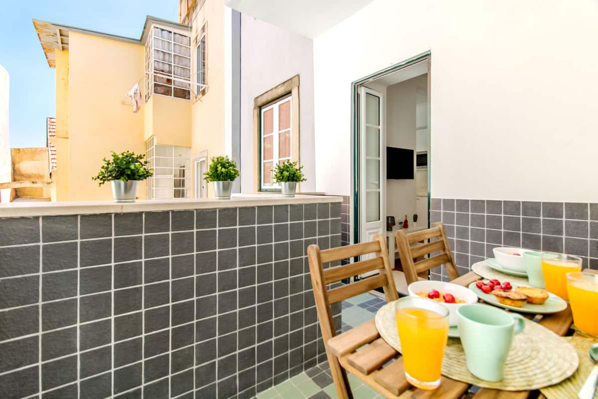 Varanda com mesa posta para café da manhã, plantas em vasos e vista do exterior do edifício.