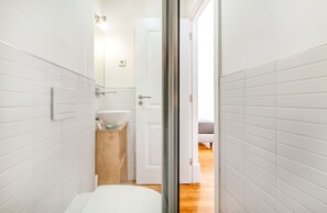 A narrow bathroom with white tiled walls, a white toilet, a wooden vanity with a glass bowl sink, and a door leading to a bedroom with a bed. - Olarias Terrace Lisbon (Lisbon)