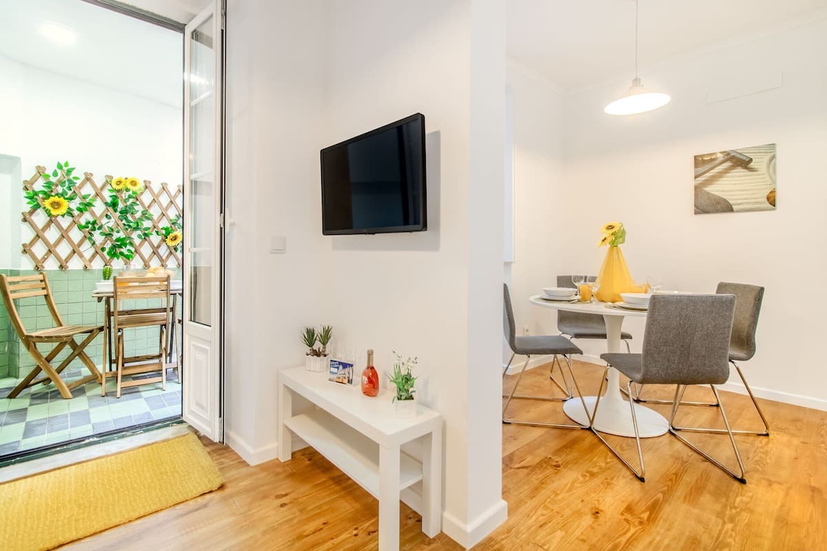 Uma sala de estar moderna com televisão, um espaço de refeições com mesa e cadeiras, e vista através de uma porta de vidro.