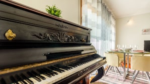 A grand piano in a room with a dining table and chairs, a plant on the piano, and a window with curtains. - São Félix Ocean Views Apartment (Vila Nova de Gaia)