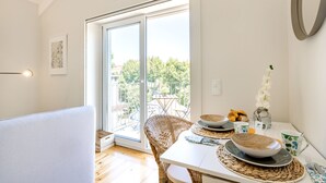 Uma área de refeições com uma mesa posta para o café da manhã, uma cadeira de vime e vista para um pátio externo através de uma porta de vidro.