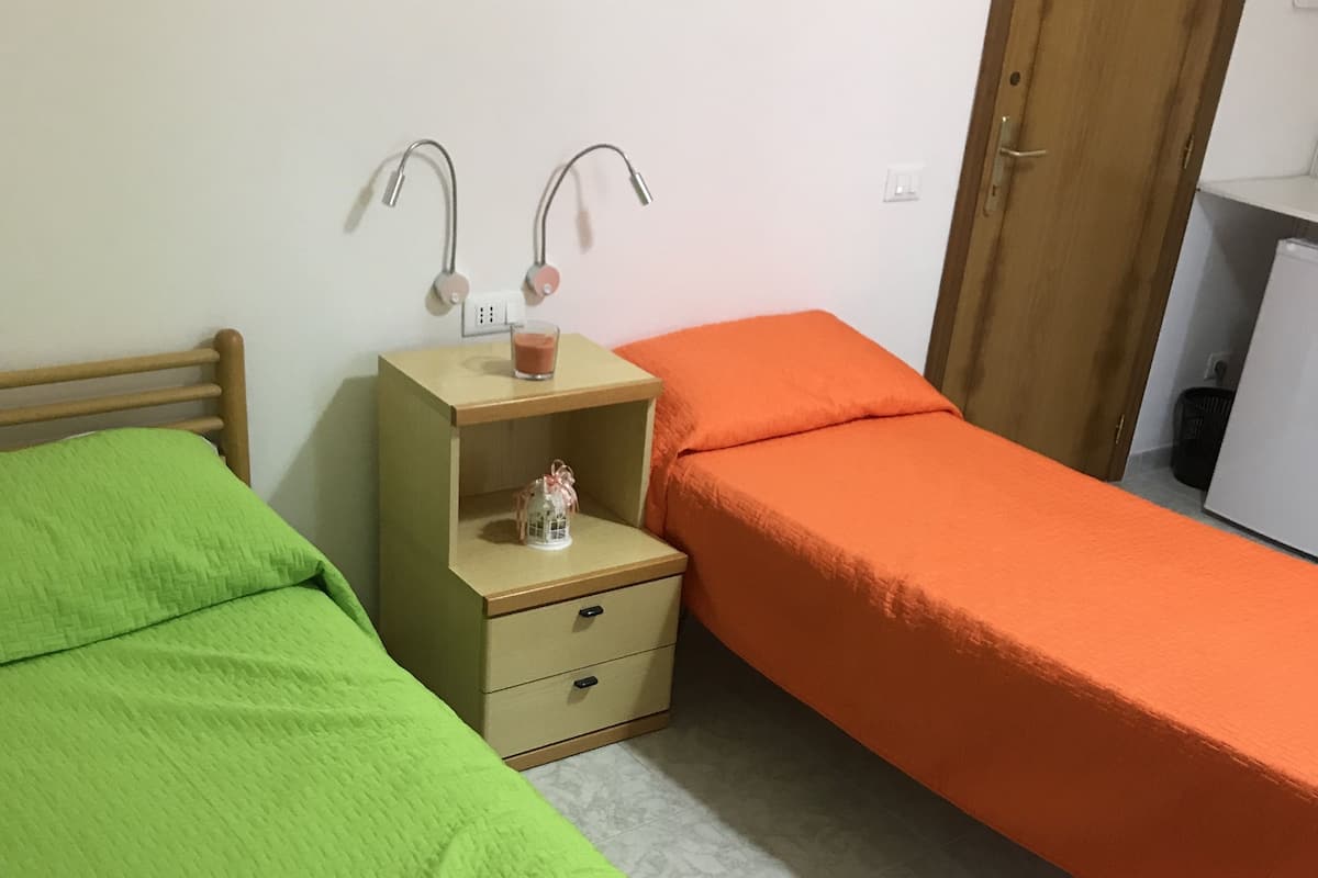 A room with two single beds, one with a green bedspread and the other with an orange bedspread. There is a wooden nightstand with a small decorative item and a drawer. A wooden door is visible in the background.