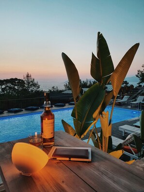 A wooden table with a bottle of liquor, a glass, and a book, overlooking a swimming pool with lounge chairs and a view of the sea and mountains. - Rocas Roja Beach Hotel (Fethiye)