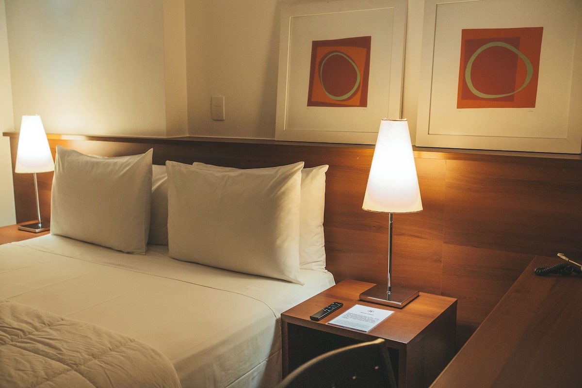 A hotel room with a bed, two bedside lamps, a nightstand with a remote control, and two framed artworks on the wall.