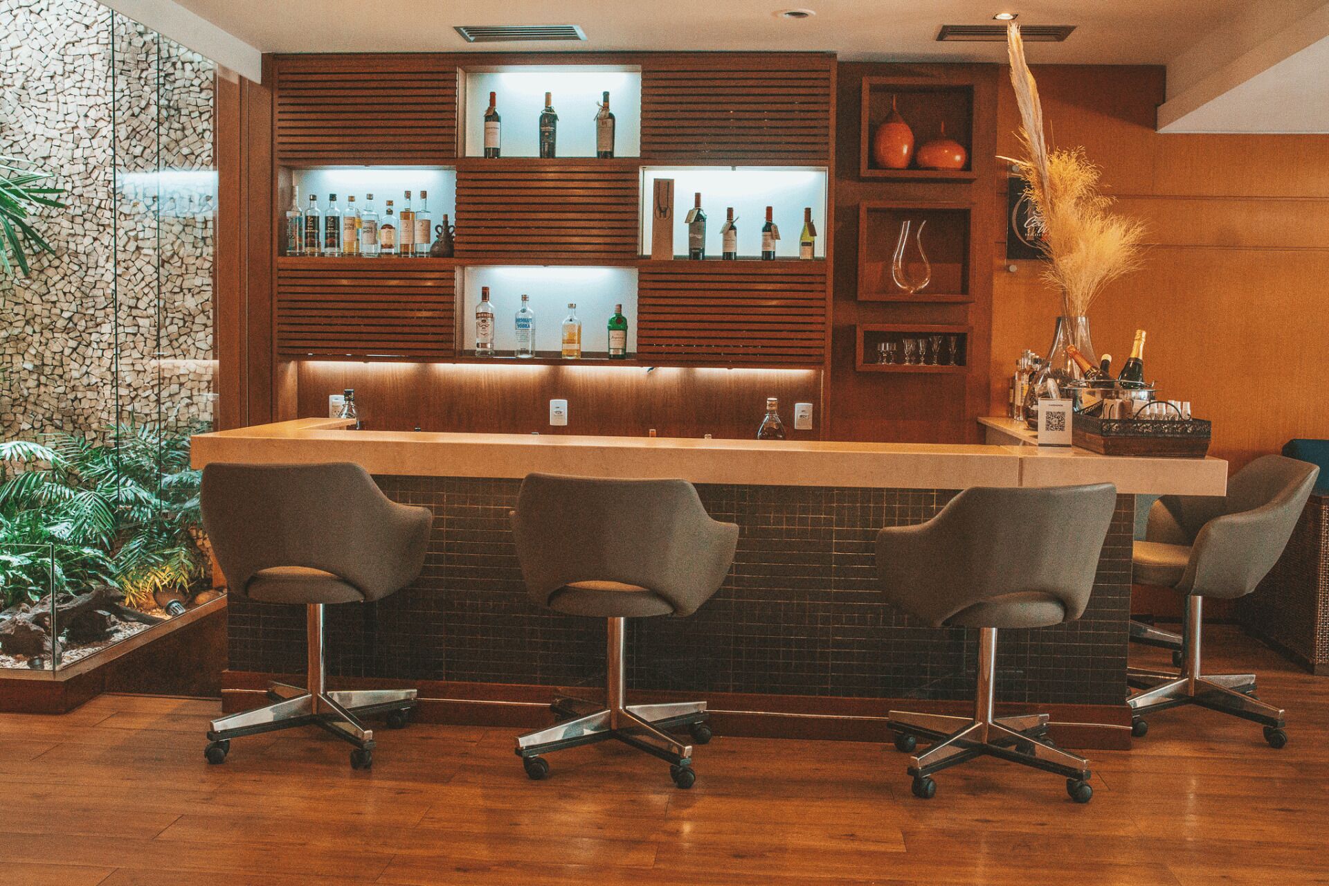 Zona de bar con mostrador de madera, tres taburetes y estantes con botellas.