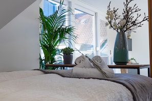 A bedroom with a bed, a potted plant, a vase with flowers, and a window with a view of buildings.
