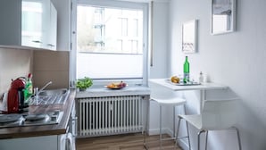Moderni keittiö, jossa on ikkuna, valkoiset kaapit, patteri, pesuallas, vedenkeitin, viherkasvi ja pieni baaritiski jakkaroineen.