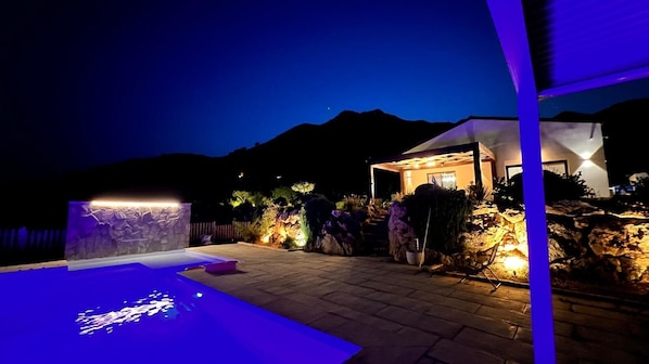 A pool area with a stone wall and a small building with a lighted entrance.