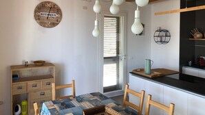 A kitchen with wooden cabinets, a dining table with chairs, hanging light bulbs, a clock, and a window with blinds.