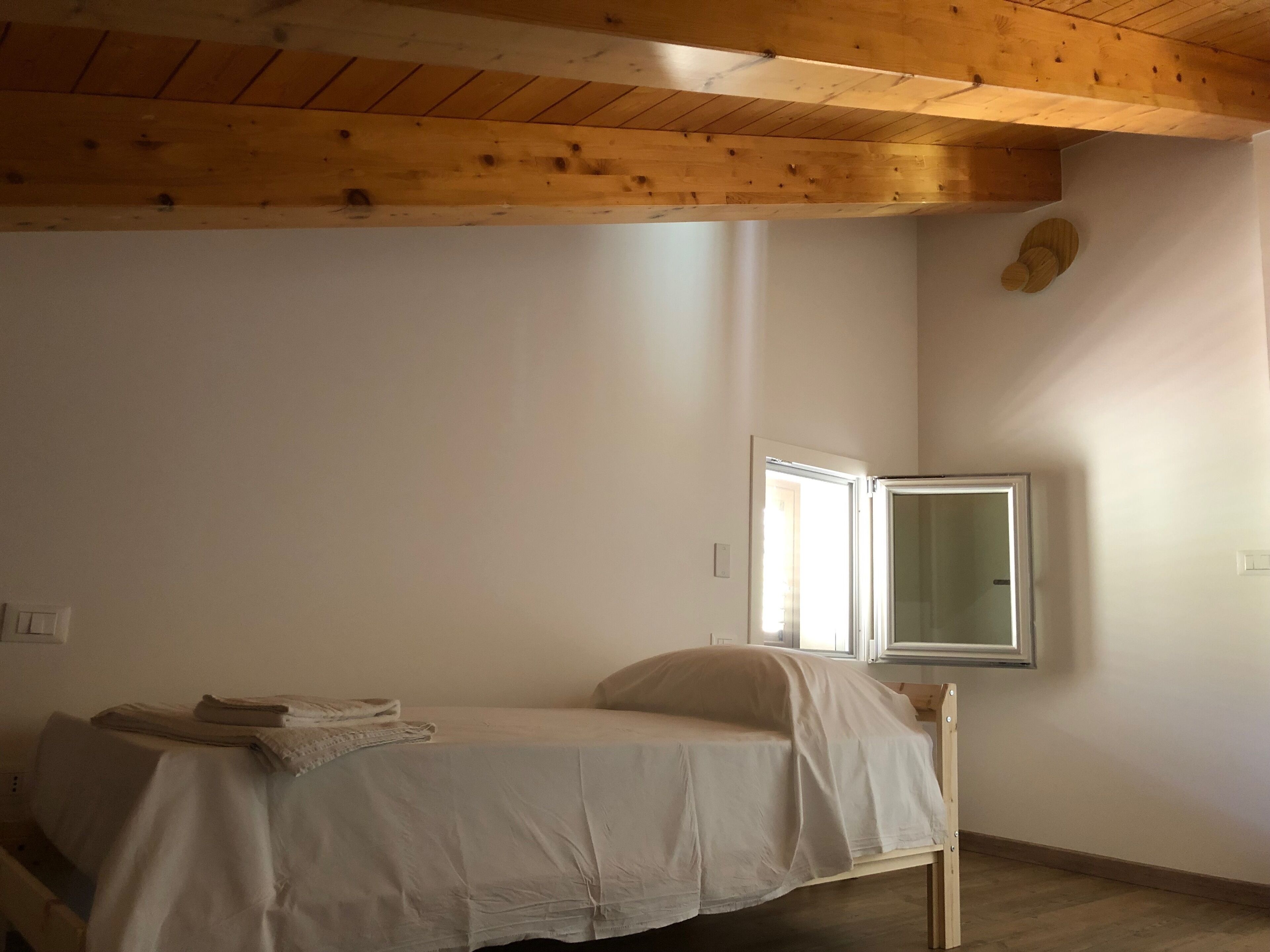 Een kleine kamer met een enkel bed, houten plafond, een raam en een spiegel aan de muur.