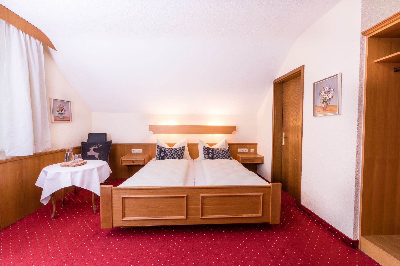 Een hotelkamer met een houten bed, twee nachtkastjes, een klein tafeltje met een witte tafelkleed en een rood tapijt.