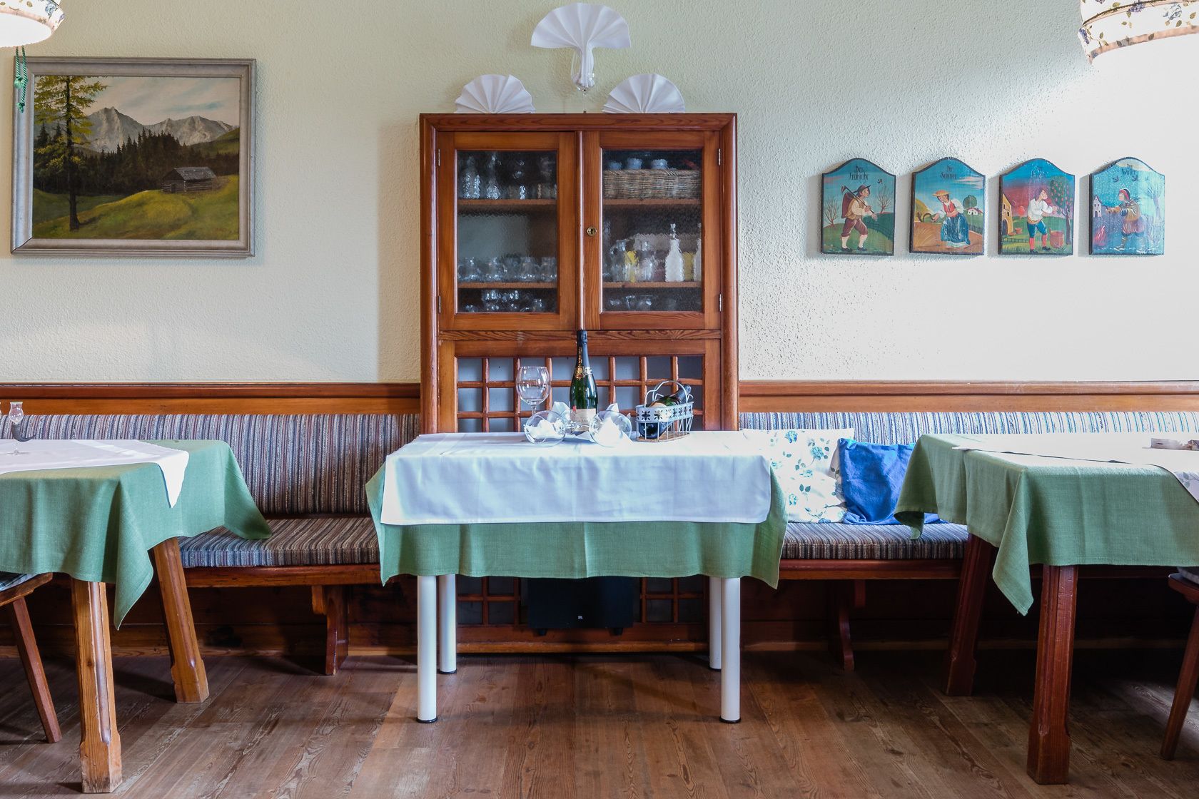 Un comedor con bancos y mesas de madera, un aparador con puertas de cristal y un cuadro en la pared.